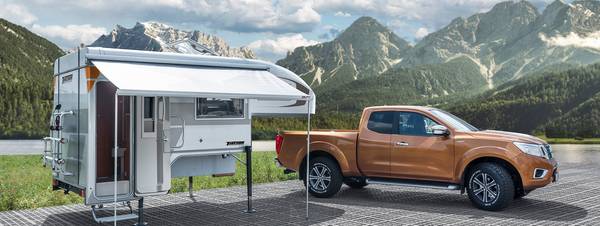 Tischer pickup camper TRAIL 260S med utfälld markis bredvid en ljusbrun Nissan Navara med höjda vikter och omtypning framför höga berg