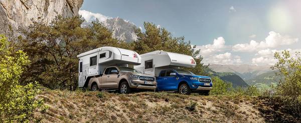 Tischer pickup camper TRAIL 260 S på en beige Ford Ranger och Tischer pickup camper Box 240 på en blå Ford Ranger stående bredvid varandra framför träd och höga berg