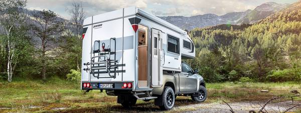 Tischer pickup camper TRAIL 260S med cykelställ och öppnad dörr på en silverfärgad Toyota Hilux stående framför skog och berg