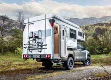 Tischer pickup camper TRAIL 260S med cykelställ och öppnad dörr på en silverfärgad Toyota Hilux stående framför skog och berg
